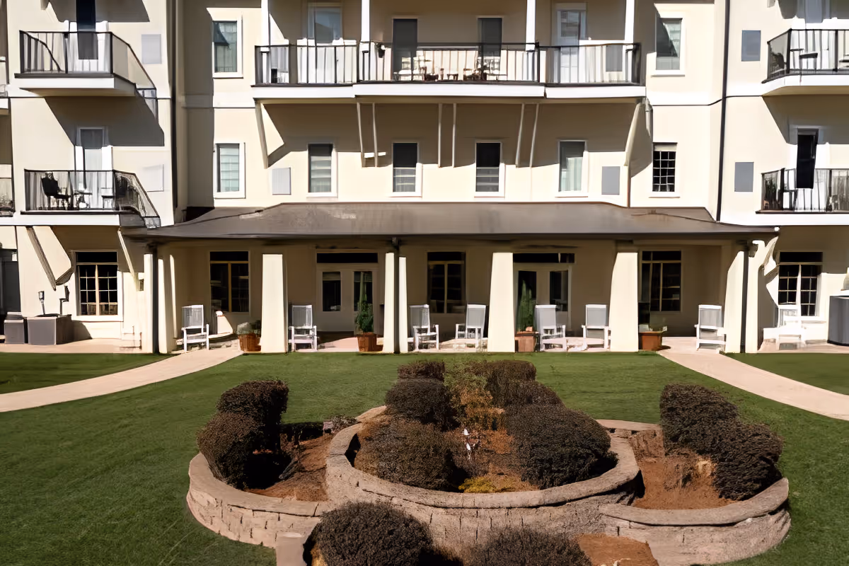 Multi-story pale yellow building with balconies, a covered patio lined with white chairs, and a manicured lawn with a raised shrub bed in the foreground.