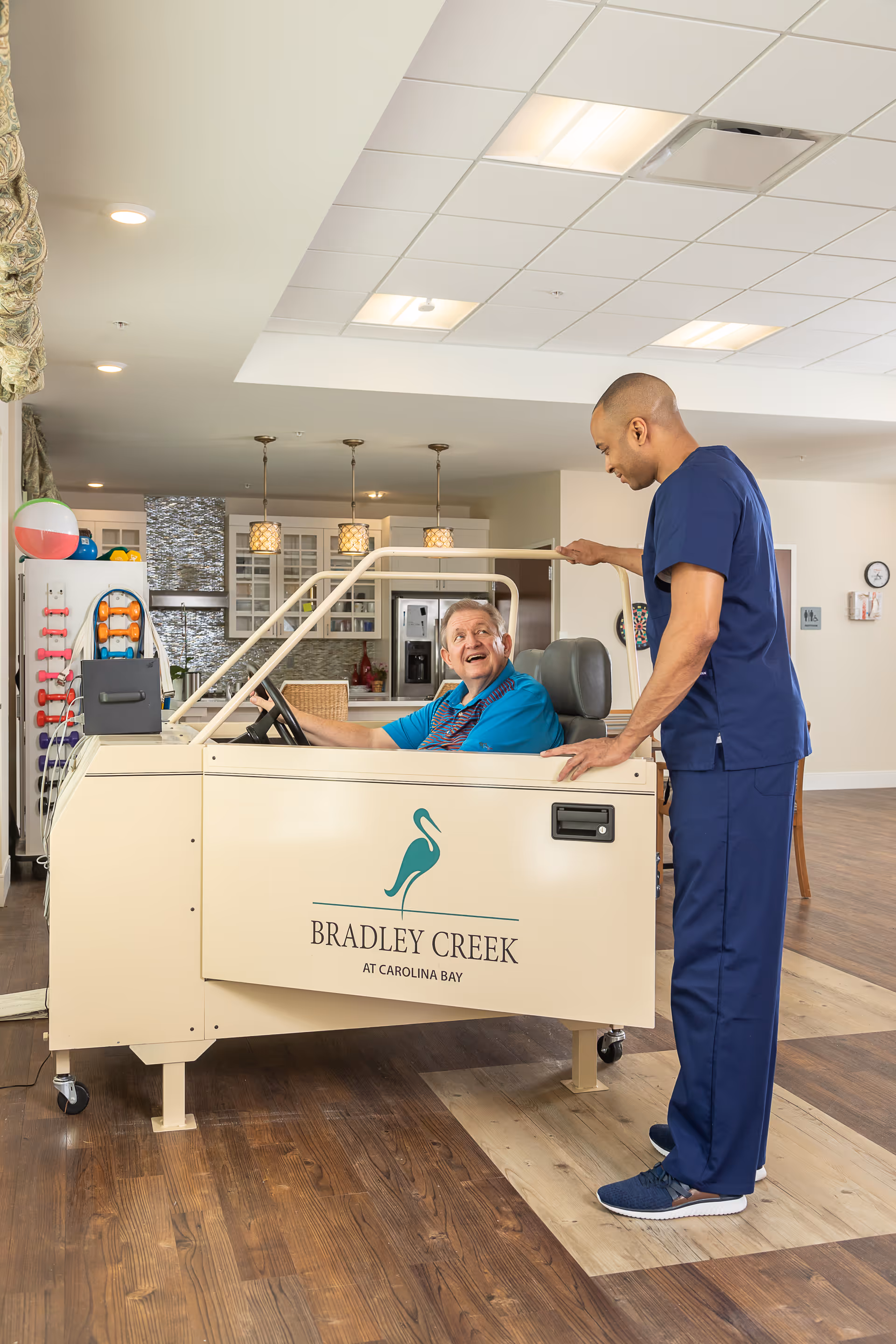 An elderly man sitting in a beige indoor driving simulator vehicle labeled 'Bradley Creek at Carolina Bay' inside a senior living facility. A caregiver in blue scrubs stands beside him, engaging in conversation. The background shows a well-lit interior with kitchen cabinets, pendant lights, and exercise equipment.