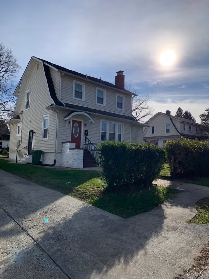 Two-story beige house with red front door, steps, and a hedge on a sunny suburban street.