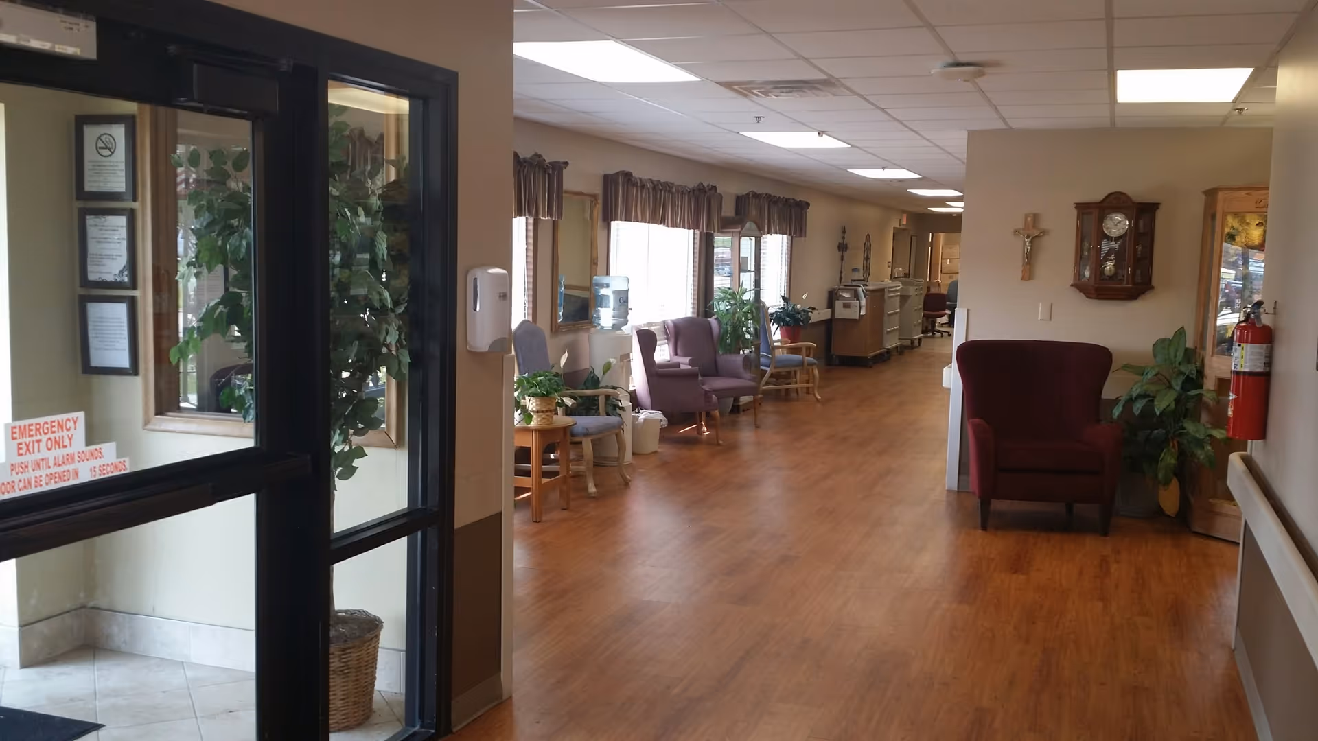 A hallway in a senior living facility with wooden flooring and beige walls. There are several chairs and plants along the wall with windows covered by valances. A maroon armchair is placed near a wall with a clock and a cross hanging above it. A fire extinguisher is mounted on the wall to the right. The entrance door with glass panels is visible on the left side.