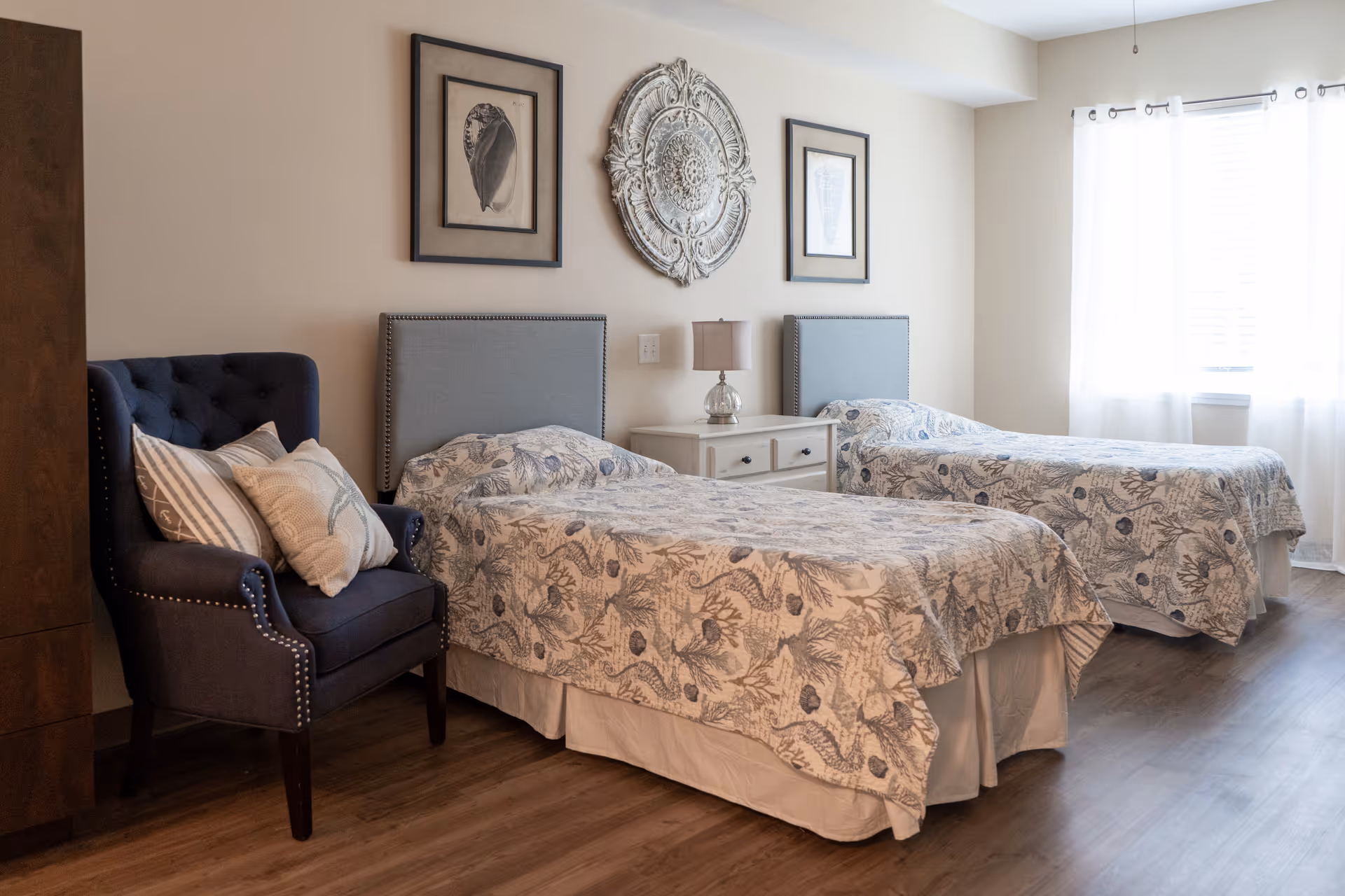 A bright bedroom with two twin beds featuring matching floral bedspreads and gray upholstered headboards. Between the beds is a white nightstand with a small lamp. On the left side of the room is a dark blue armchair with decorative pillows. The walls are decorated with two framed seashell prints and a large ornate circular wall decoration. Light filters in through a window with sheer white curtains on the right.