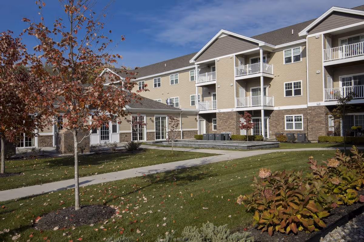 Three-story beige senior living building with balconies overlooking a landscaped courtyard, sidewalks, and autumn trees.