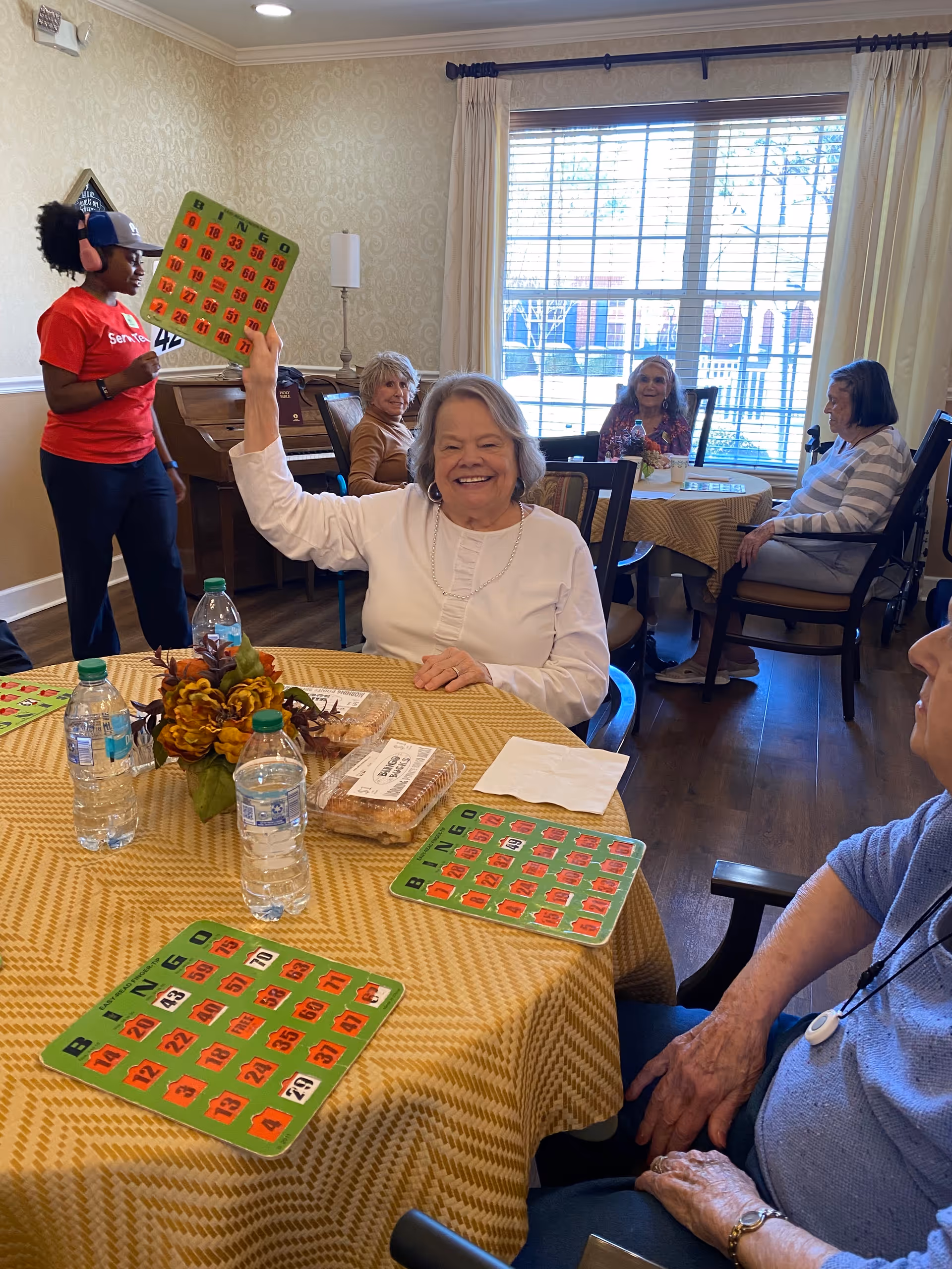 A group of elderly women sitting around tables playing bingo in a well-lit room with large windows. One woman in a white shirt is smiling and holding up her bingo card. A staff member wearing a red shirt and headphones stands nearby. The tables are covered with yellow patterned tablecloths and have water bottles, snacks, and a flower arrangement on them.