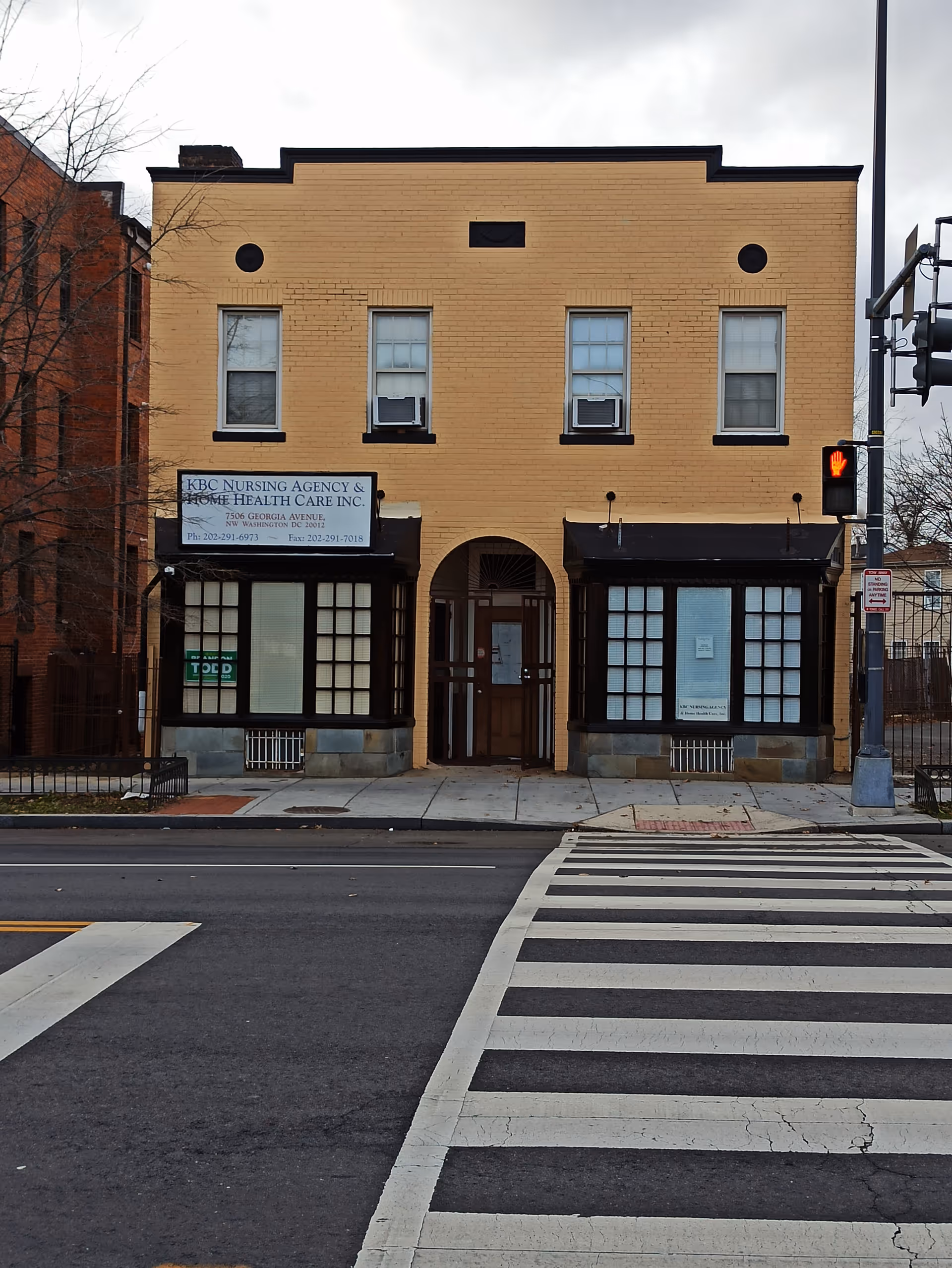 Two-story tan brick building with storefronts and a sign for KBC Nursing Agency on a street.