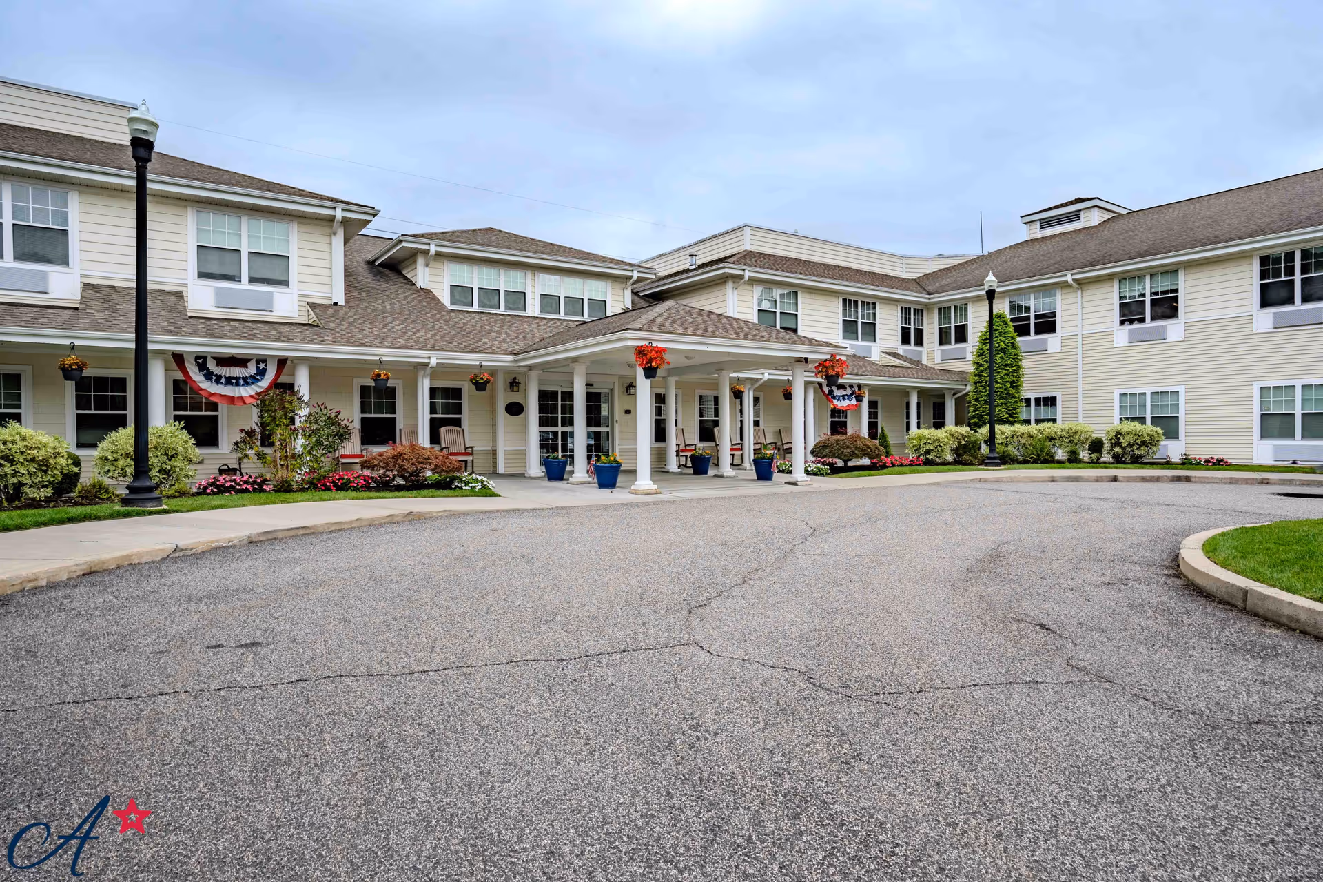 Front exterior view of a two-story assisted living facility with beige siding, multiple windows, a covered entrance with white columns, hanging flower baskets, and a circular driveway.