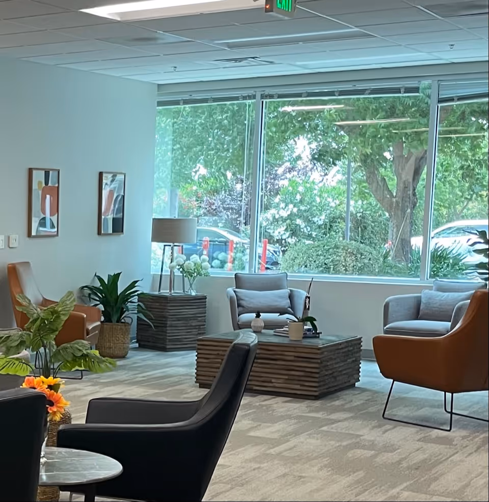 Bright lounge area with modern chairs, a wooden coffee table, plants, and large windows.