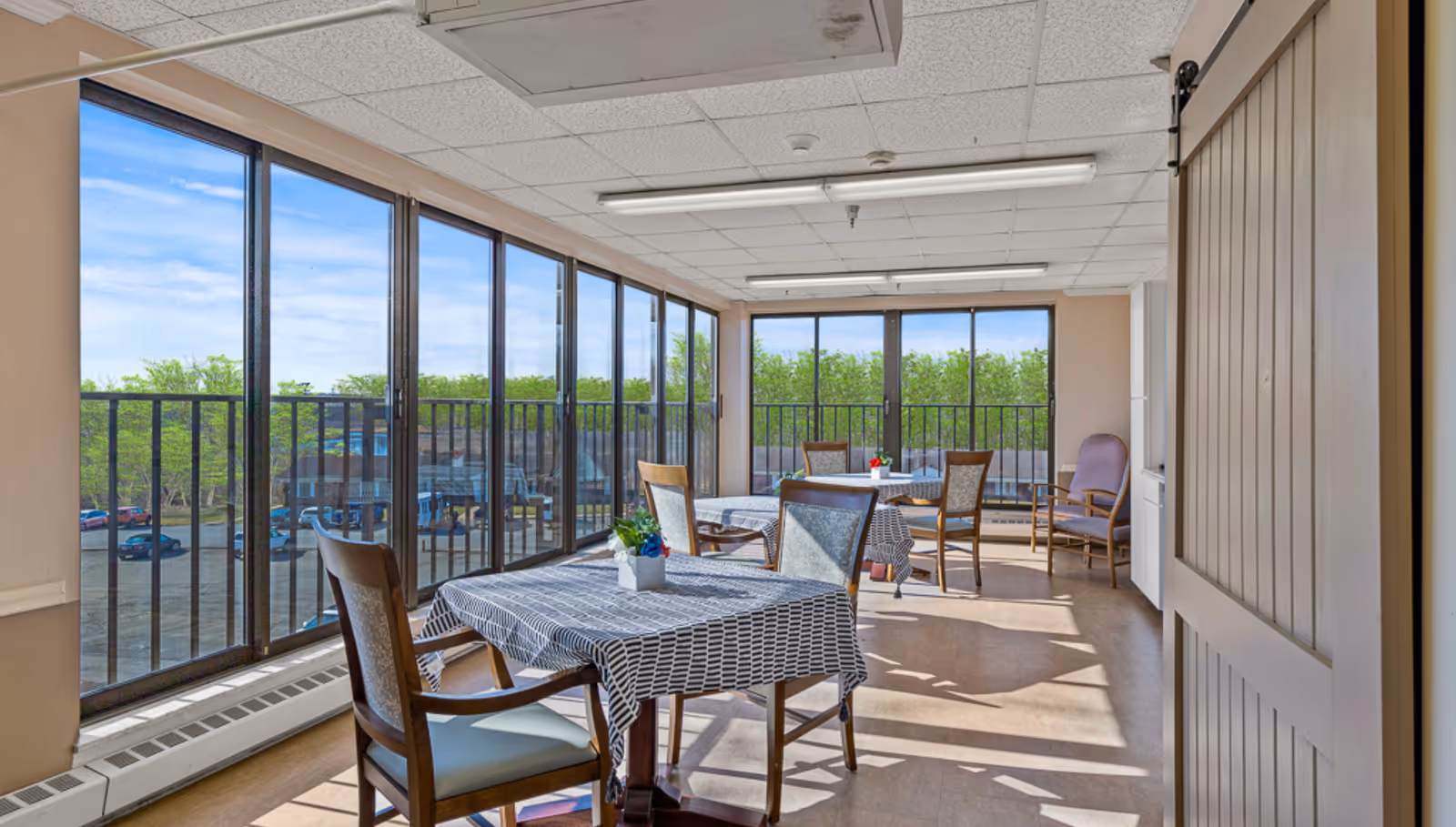 Sunlit dining area with tables, chairs, and patterned tablecloths by large windows.