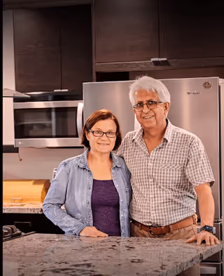 An elderly couple standing together in a modern kitchen with dark cabinets, a stainless steel refrigerator, and a microwave. They are smiling and standing behind a granite countertop.