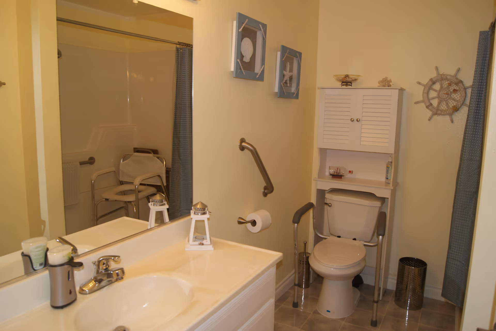 A bathroom with a sink and countertop on the left, a large mirror above the sink, a toilet with safety rails and a white cabinet above it, a shower area with a chair and grab bars, and nautical-themed wall decorations.