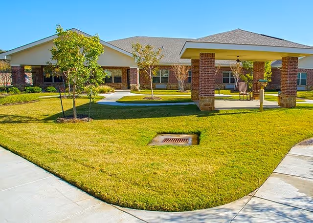 Brick single-story senior living building with a covered patio, green lawn, young trees, and paved walkways in a courtyard.