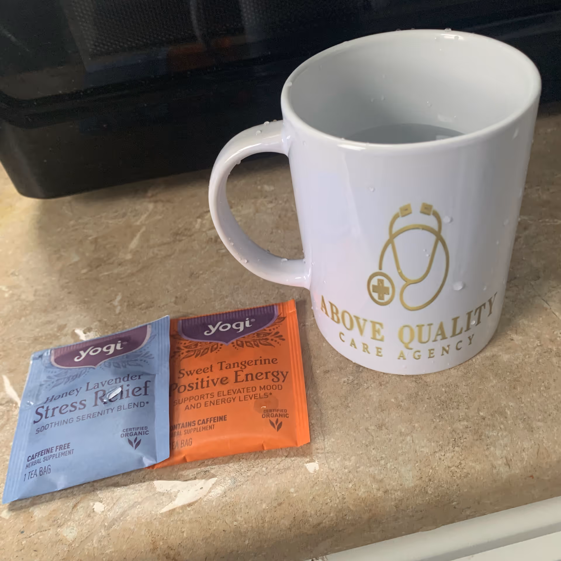 Close-up of a white mug with Above Quality Care Agency logo beside tea sachets.