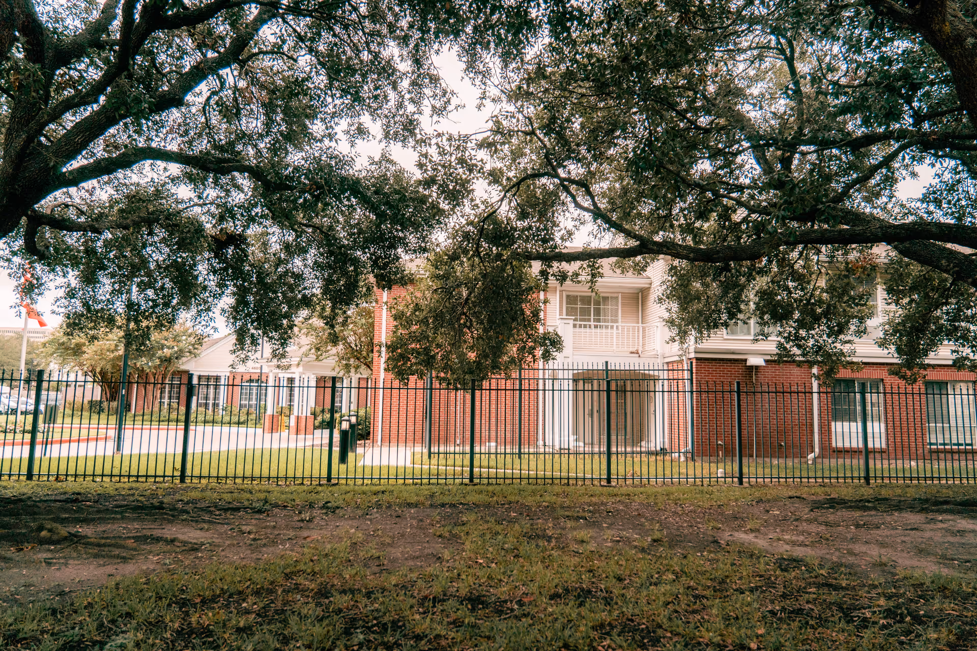 Brick two-story residential building behind a metal fence framed by large tree branches and a grassy lawn.