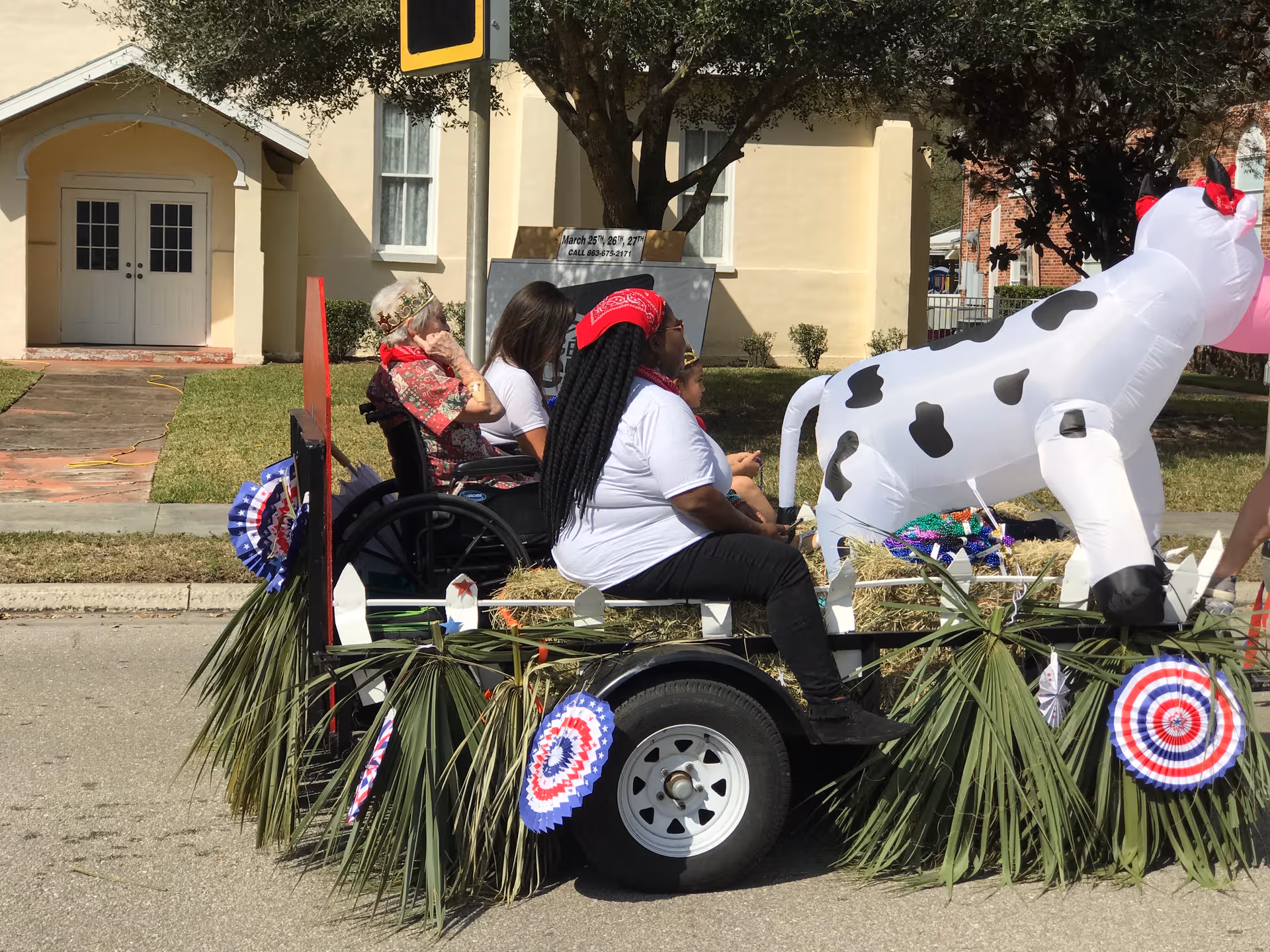 Residents ride a decorated parade float outdoors, featuring a white cow inflatable.
