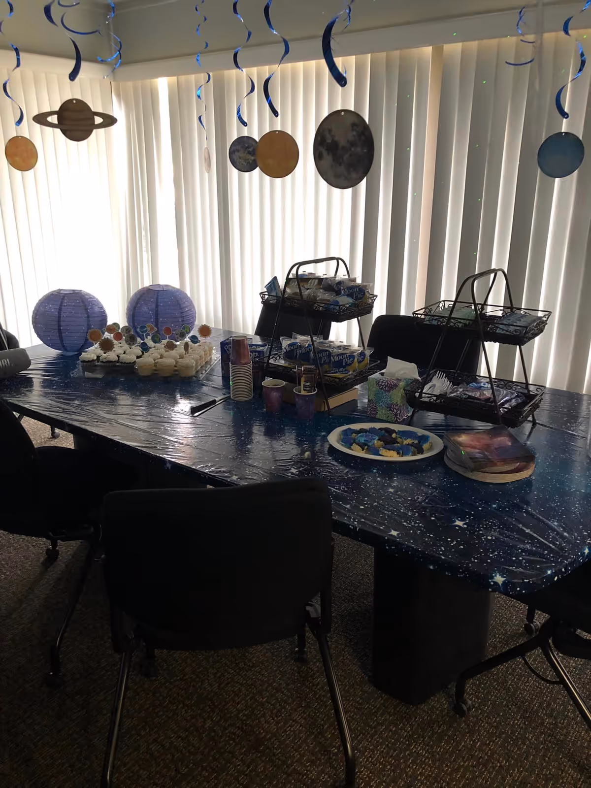 Indoor dining table decorated with space-themed decorations, cupcakes, and snacks.
