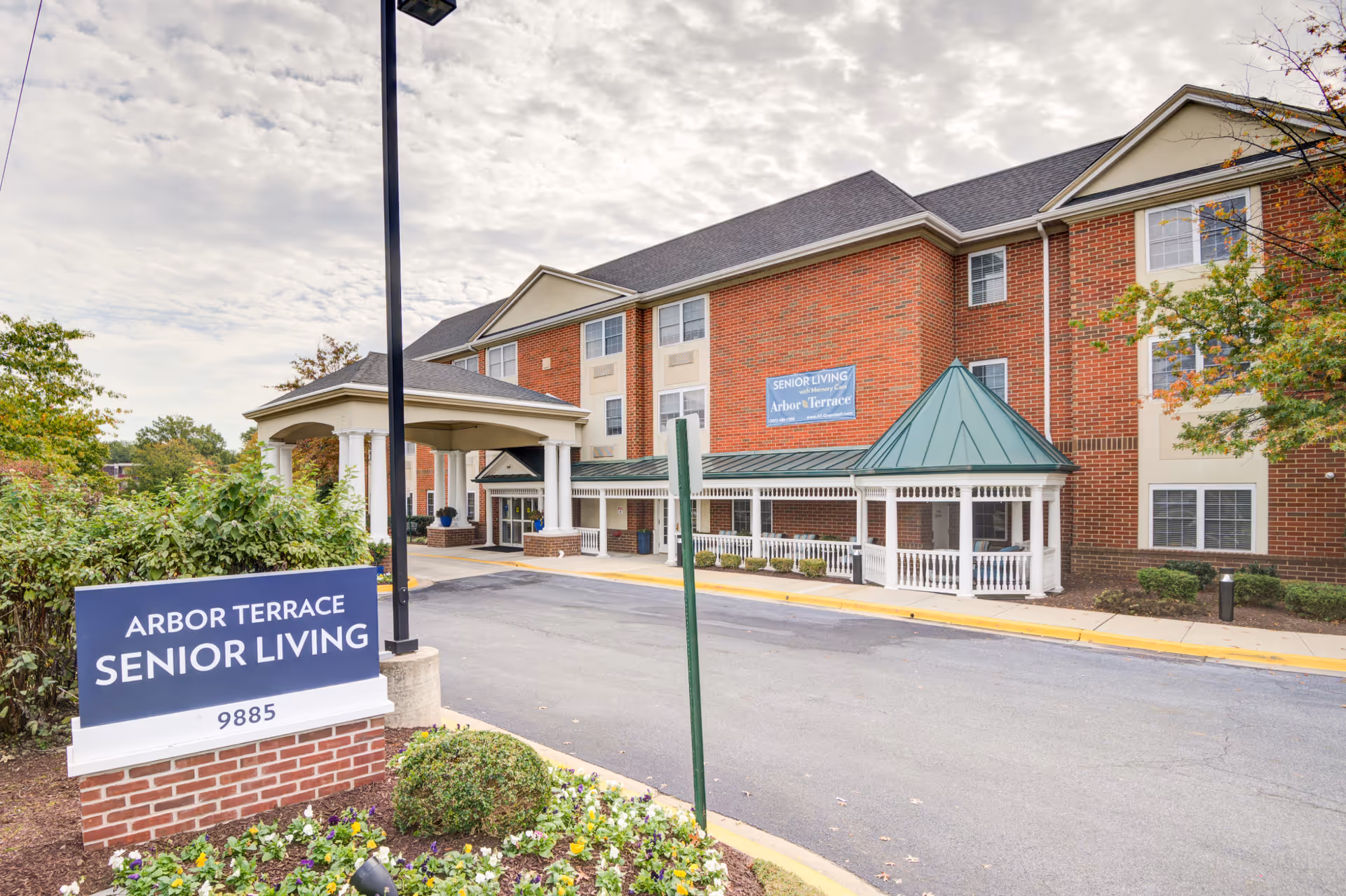 Exterior view of Arbor Terrace Senior Living facility showing a three-story brick building with white trim and a green roofed entrance canopy. There is a sign in the foreground that reads 'Arbor Terrace Senior Living 9885' surrounded by landscaping and flowers. The sky is partly cloudy.