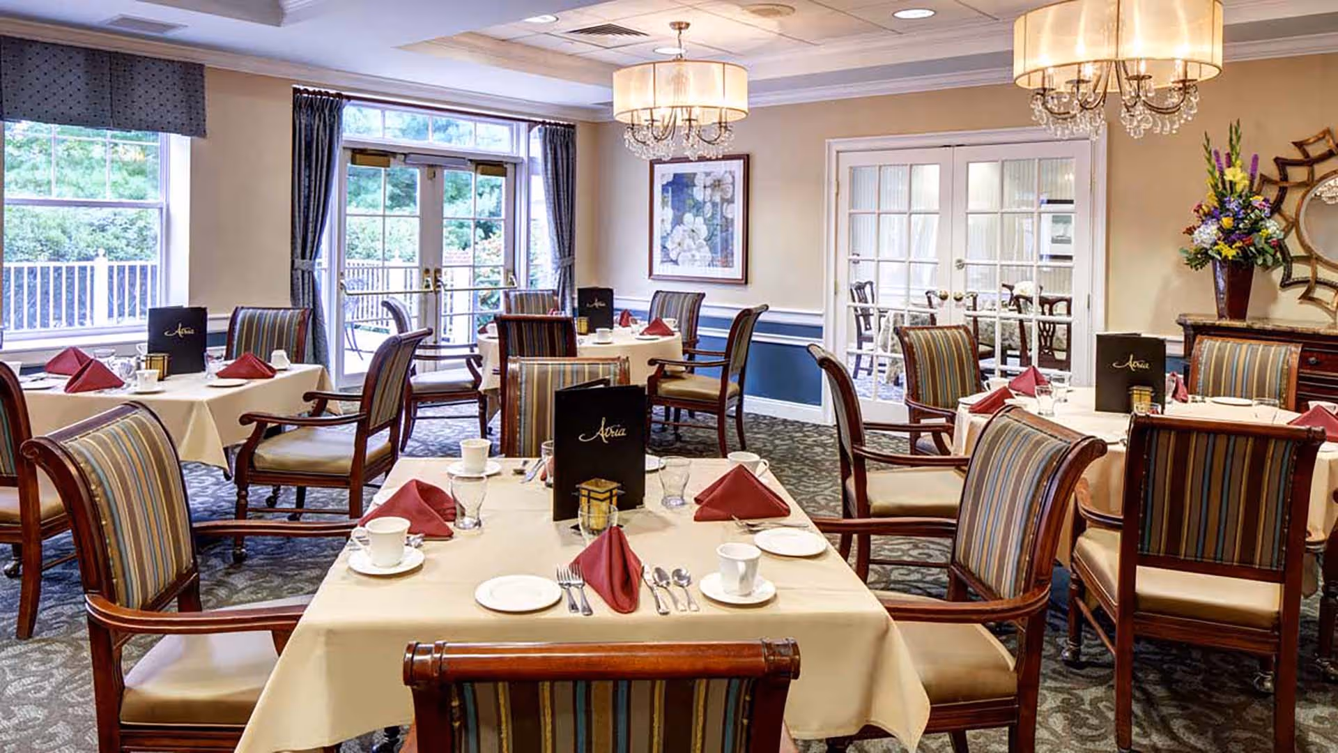 A well-lit dining room with several tables covered in beige tablecloths, each set with white cups, plates, silverware, and folded burgundy napkins. The chairs have wooden frames with striped upholstery. Large windows and glass doors let in natural light, and elegant chandeliers hang from the ceiling. There is a floral arrangement on a side table and framed artwork on the wall.