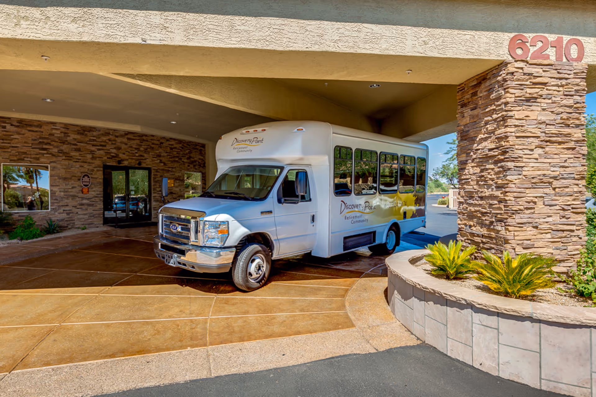 A white shuttle bus parked under a covered entrance of a building with stone pillars and the number 6210 displayed on the pillar. The bus has the text 'Discovery Point Retirement Community' on its side. There are plants in a raised stone planter near the entrance.
