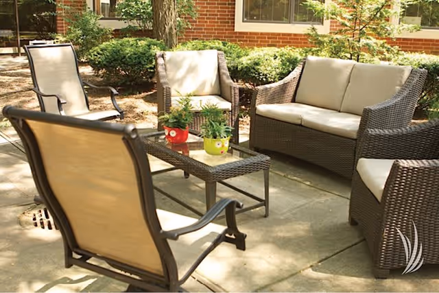 Outdoor patio area with cushioned wicker chairs and a loveseat arranged around a glass-top coffee table with two small potted plants. The patio is surrounded by greenery and a brick building with windows.