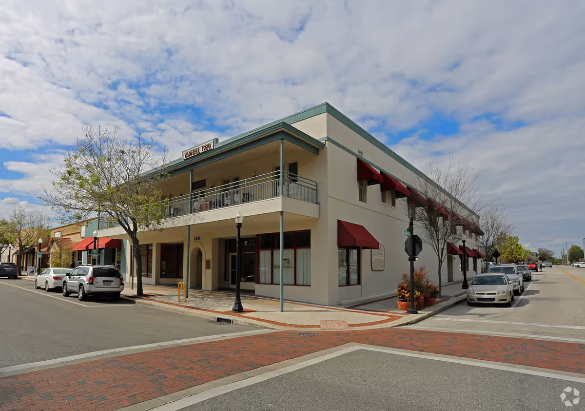 Two-story corner building with red awnings on a brick-paved street, parked cars along the curb.