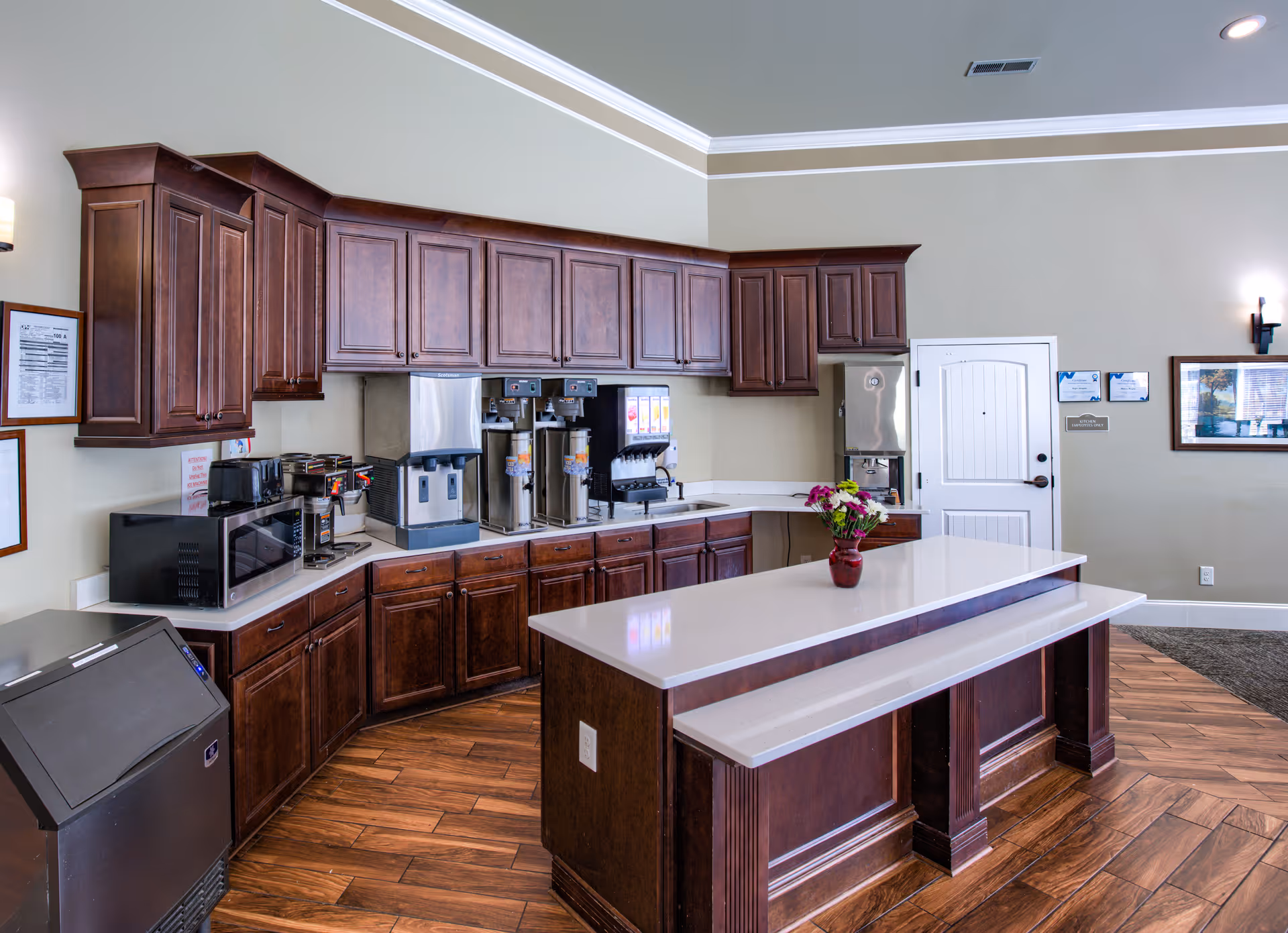 A clean and modern kitchen area with dark wooden cabinets, a white countertop island with a vase of flowers, coffee machines, a microwave, and a beverage dispenser. The room has wood-patterned flooring and light-colored walls with crown molding.