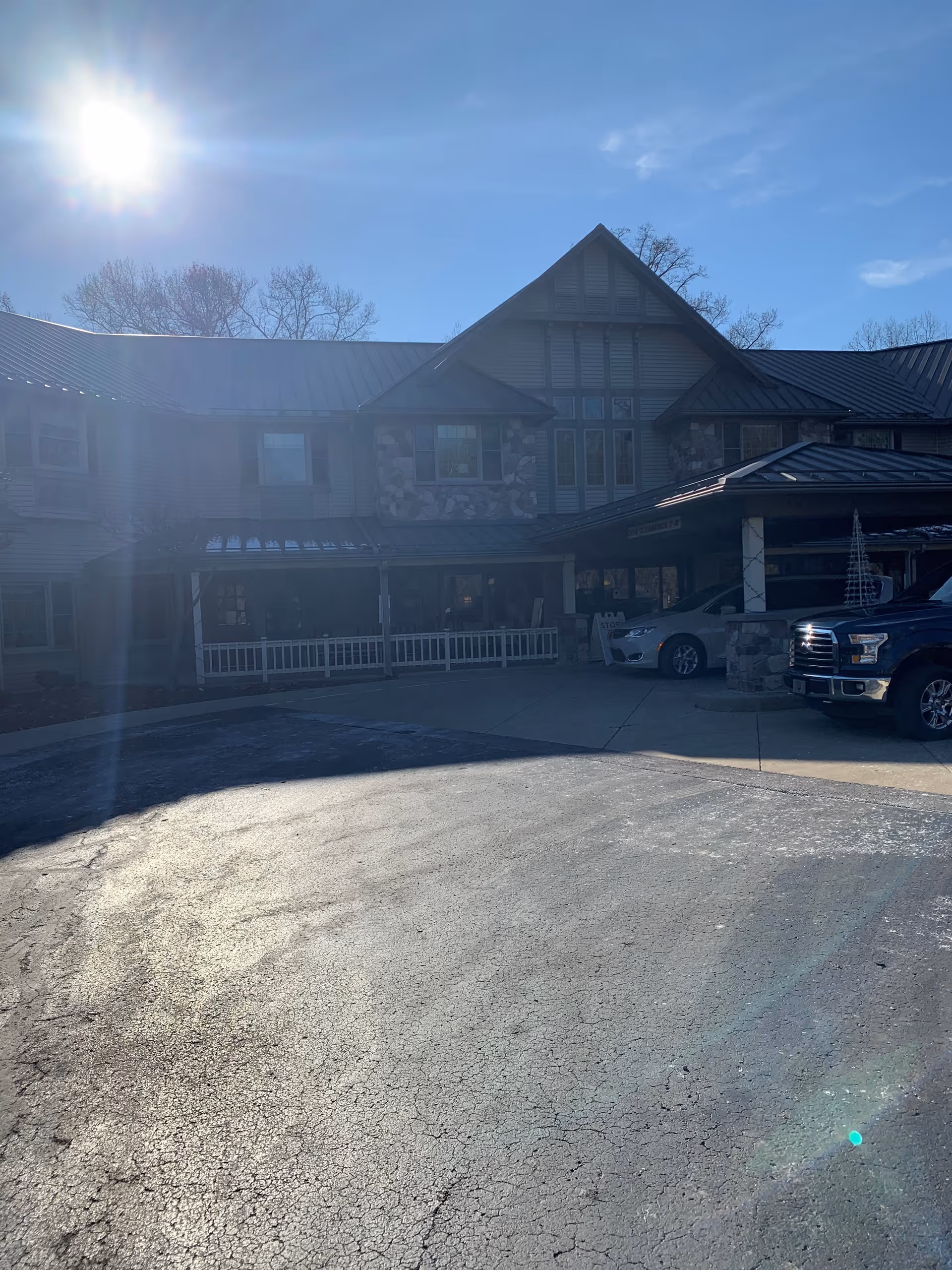 Front exterior of a two-story senior living building with a covered entrance, parked cars, and bright sun glare.