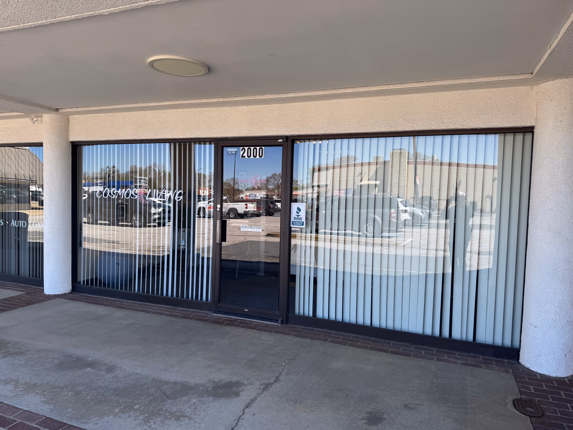 Cosmos Living storefront entrance with glass doors and vertical blinds, reflection of cars outside.