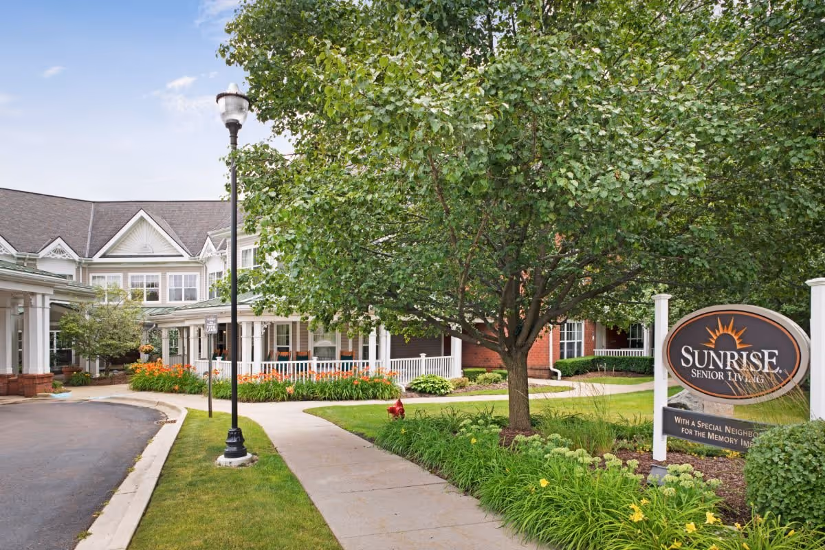 Front entrance of Sunrise Senior Living with a landscaped walkway, trees, and a large facility sign.