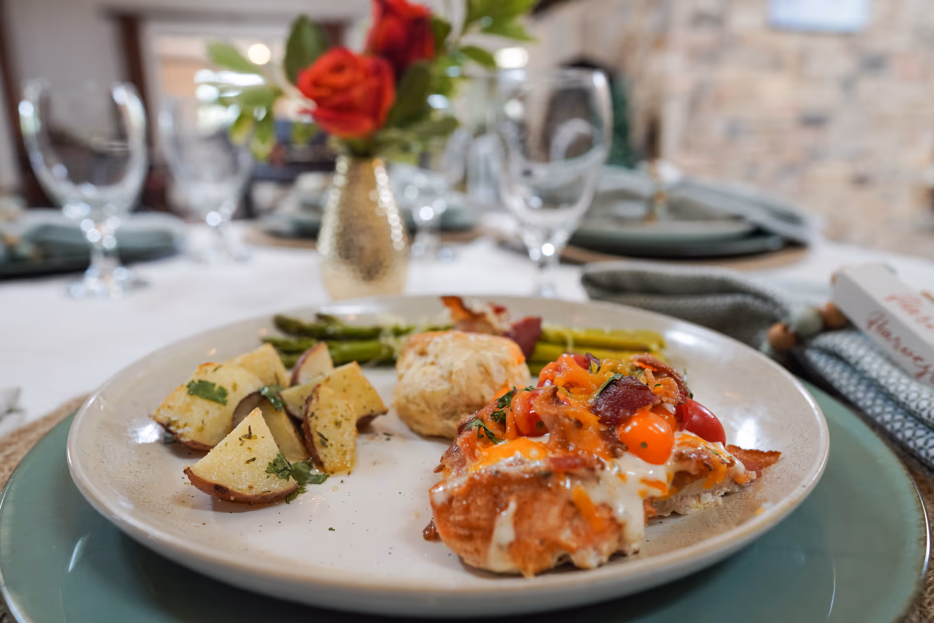 A plated meal of roasted potatoes, asparagus, a biscuit, and cheesy topped chicken sits on a set dining table with wine glasses and a small vase of red flowers in the background.