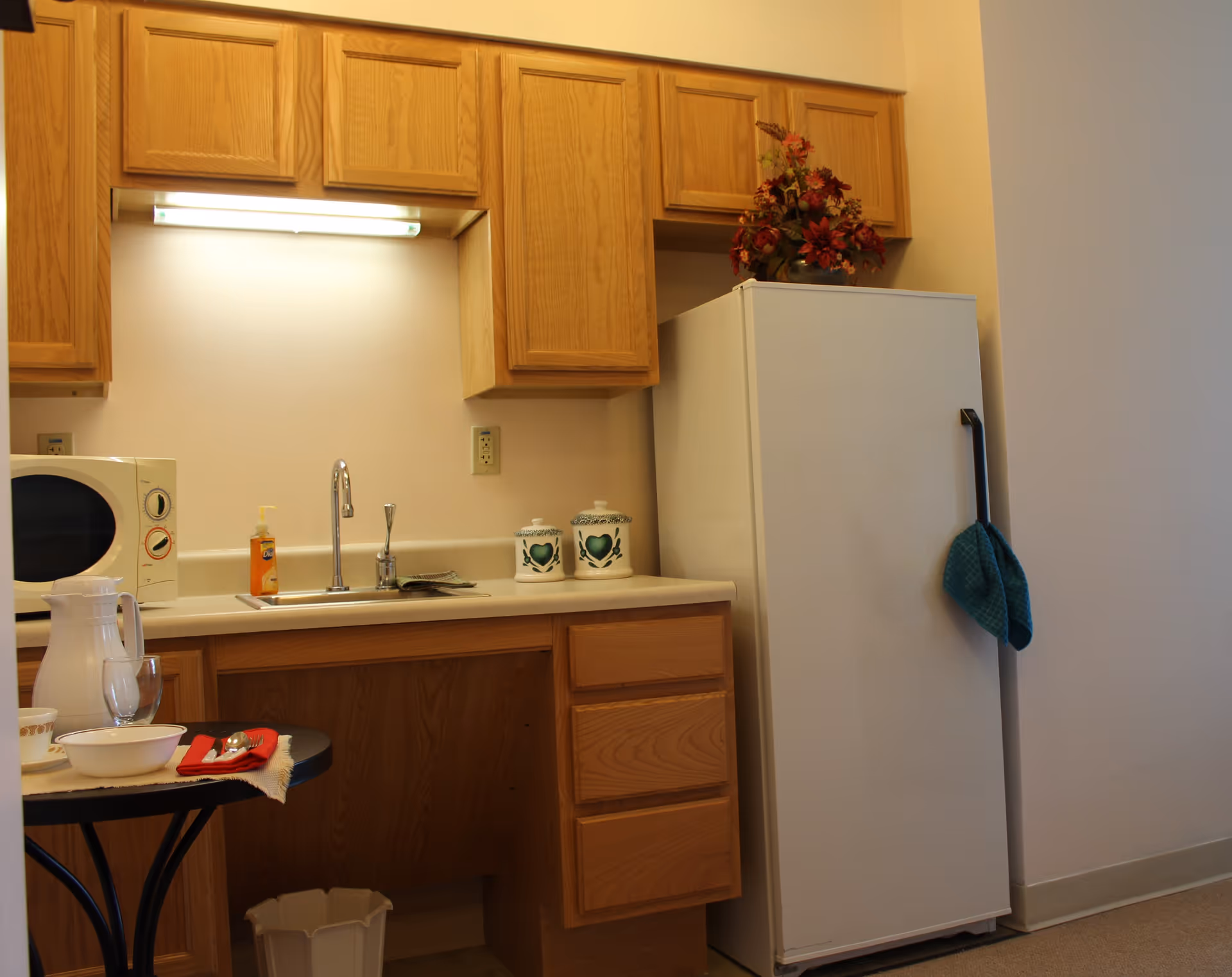 Small kitchen area with wooden cabinets, a white refrigerator with a blue towel hanging on the handle, a microwave, a sink with a faucet, soap dispenser, two decorative canisters, and a small round table set with a pitcher, glass, bowl, and utensils on a red napkin.