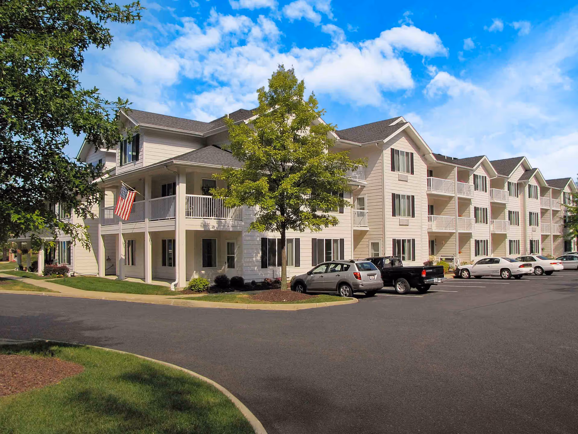 Exterior view of a three-story beige senior living building with balconies, a parking lot, trees, and an American flag.