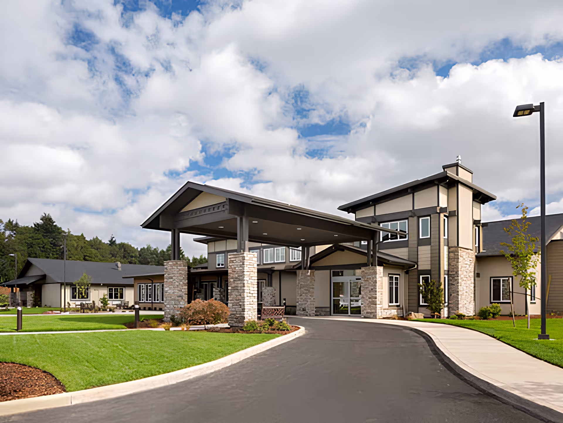 Entrance of a modern senior living building with a covered drop-off canopy supported by stone columns, paved drive, and manicured lawns under a partly cloudy sky.