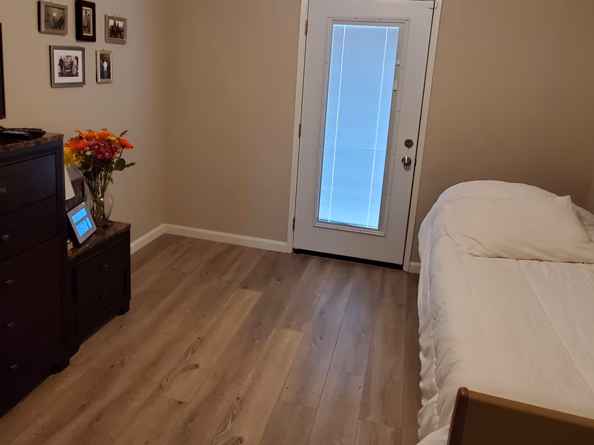 A bedroom with light wood flooring, a bed with white bedding on the right, a dark wooden dresser and nightstand on the left with a vase of colorful flowers and framed photos on the wall. A door with a window covered by blinds is centered in the background.