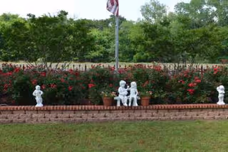 A garden area with a brick border featuring small white statues of children and potted plants with red flowers. An American flag on a pole is visible in the background with green trees and bushes behind the garden.