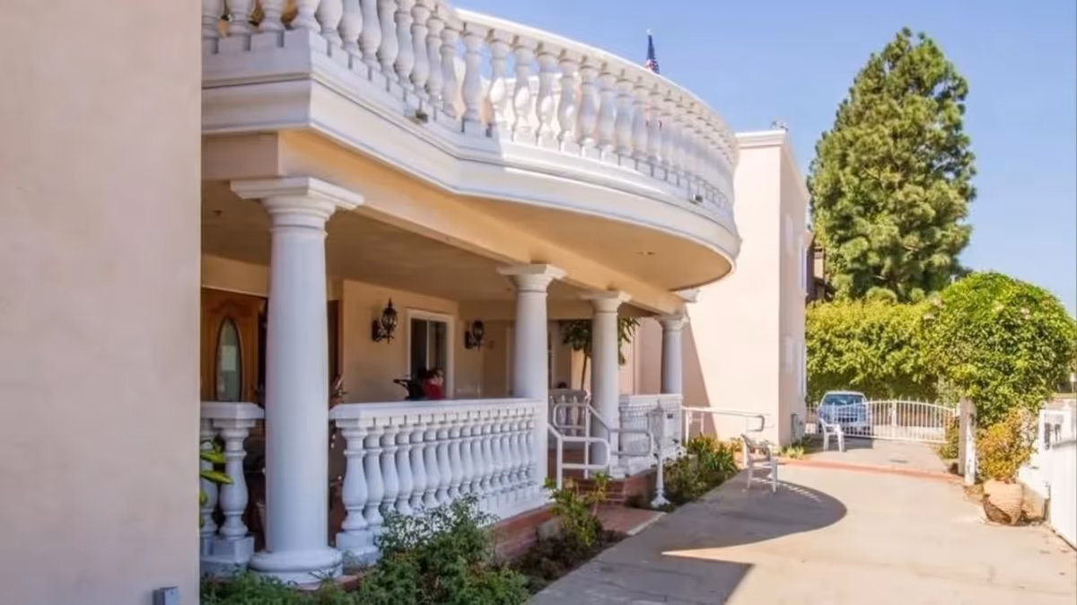 Front exterior of a two-story building with white balustrade balconies, columns, a driveway and landscaping.