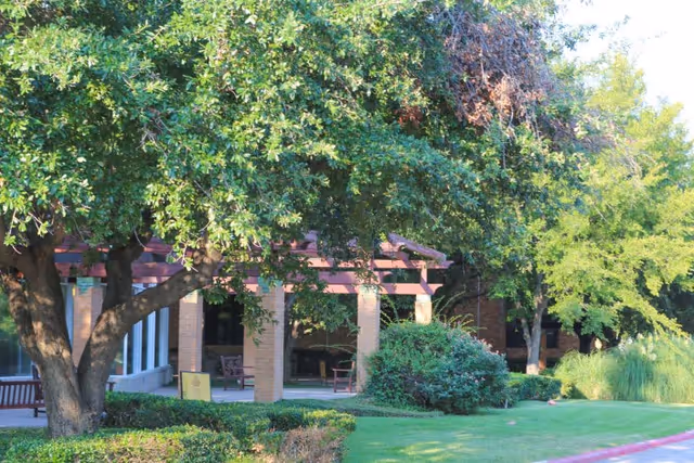 Shaded brick patio with a pergola, benches, and trees in front of the senior living facility.