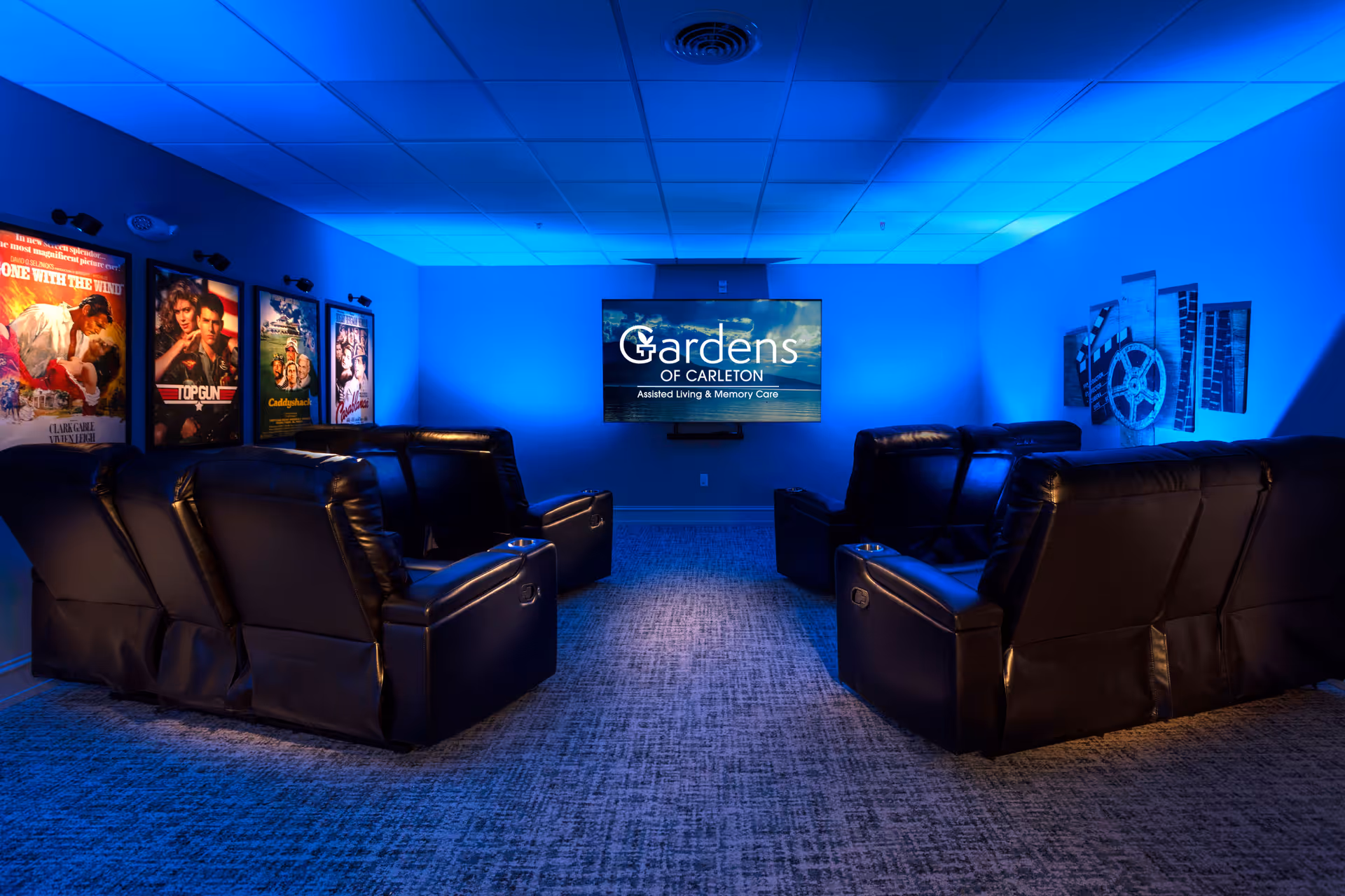 A cozy movie theater room with blue ambient lighting, featuring two rows of black leather recliner chairs facing a large flat-screen TV displaying the Gardens of Carleton logo. Movie posters are mounted on the left wall, and a film reel decoration is on the right wall.
