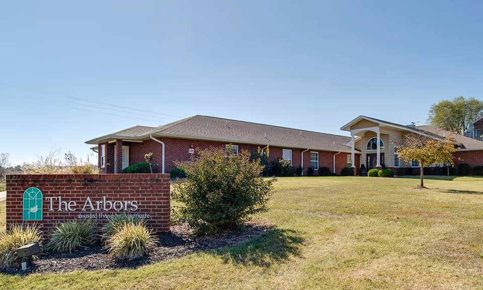 Brick single-story senior living building with a lawn and a brick sign reading "The Arbors" in the foreground.