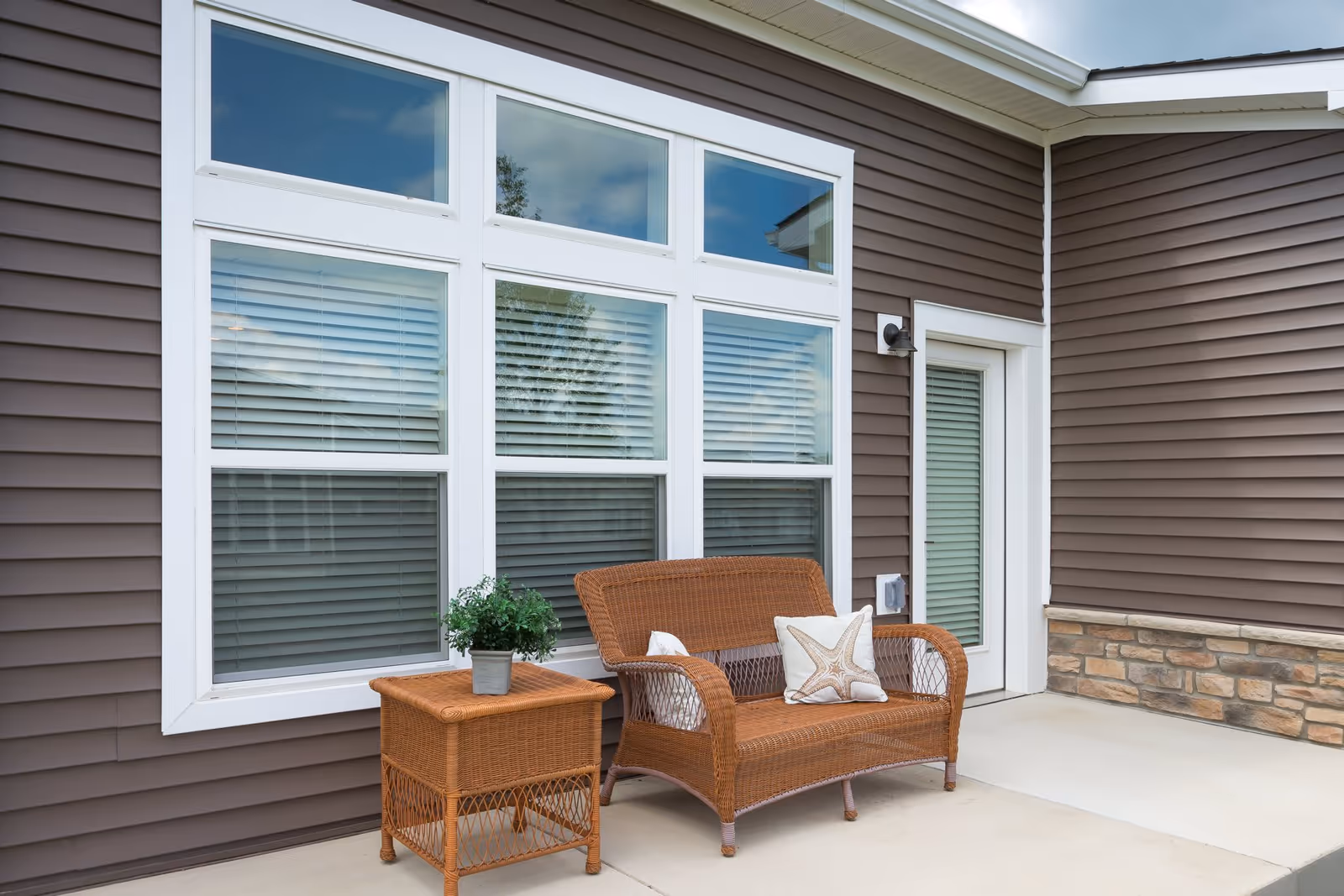 Wicker patio bench and side table with a potted plant and cushion on a concrete porch in front of large white-framed windows and brown siding.