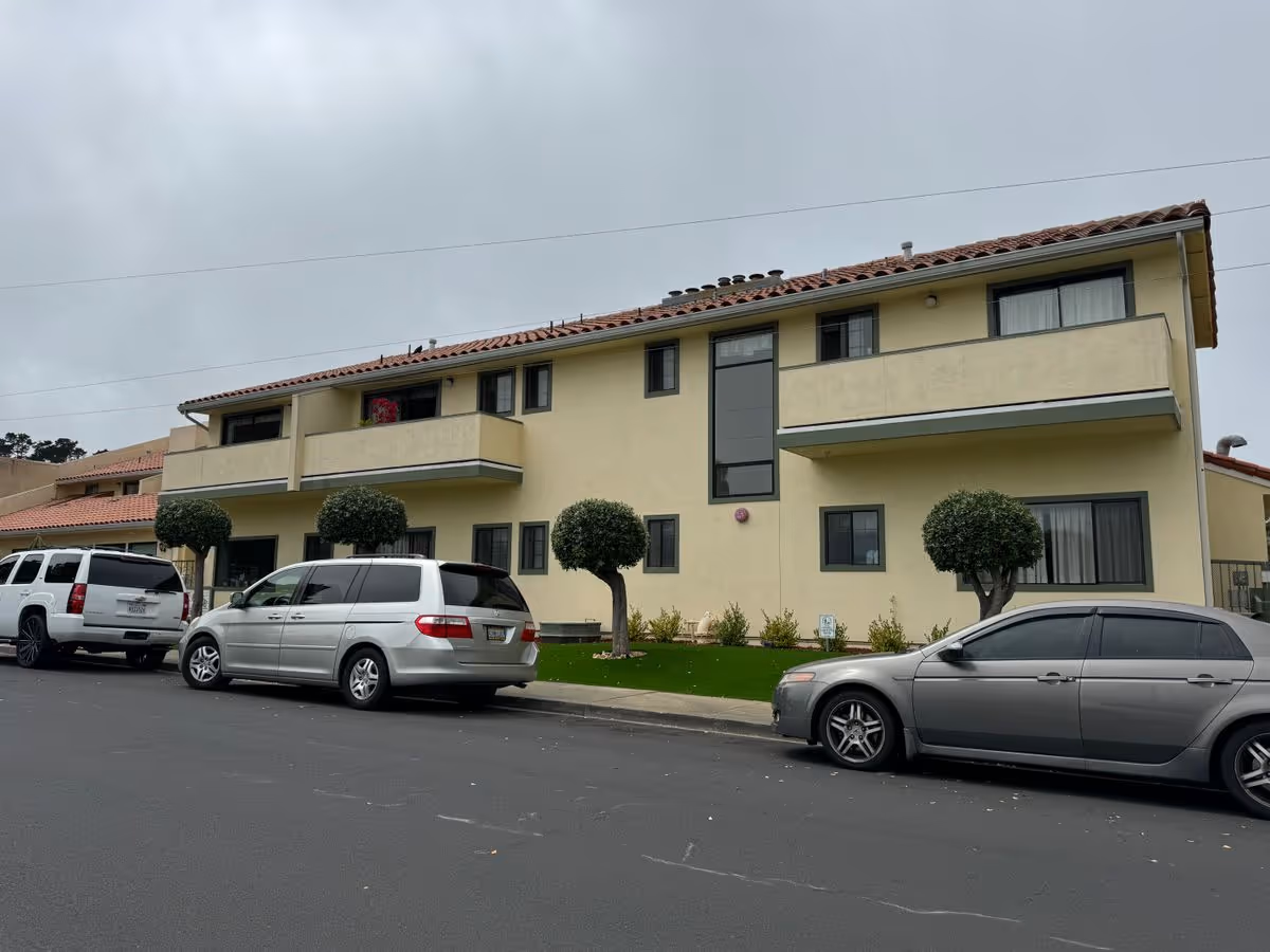 Two-story yellow stucco building with balconies, trimmed round trees and several cars parked on the street in front.