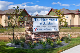 Front exterior of a senior living campus with a stone sign reading The Homestead at Anoka