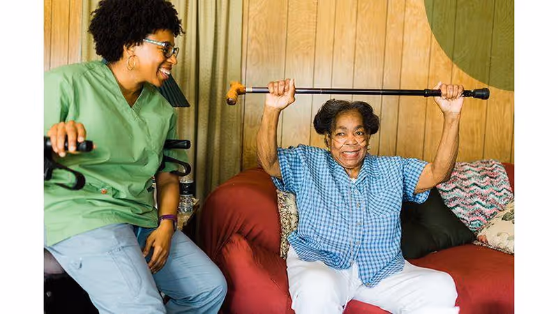 Caregiver and resident exercise together indoors in a living room.