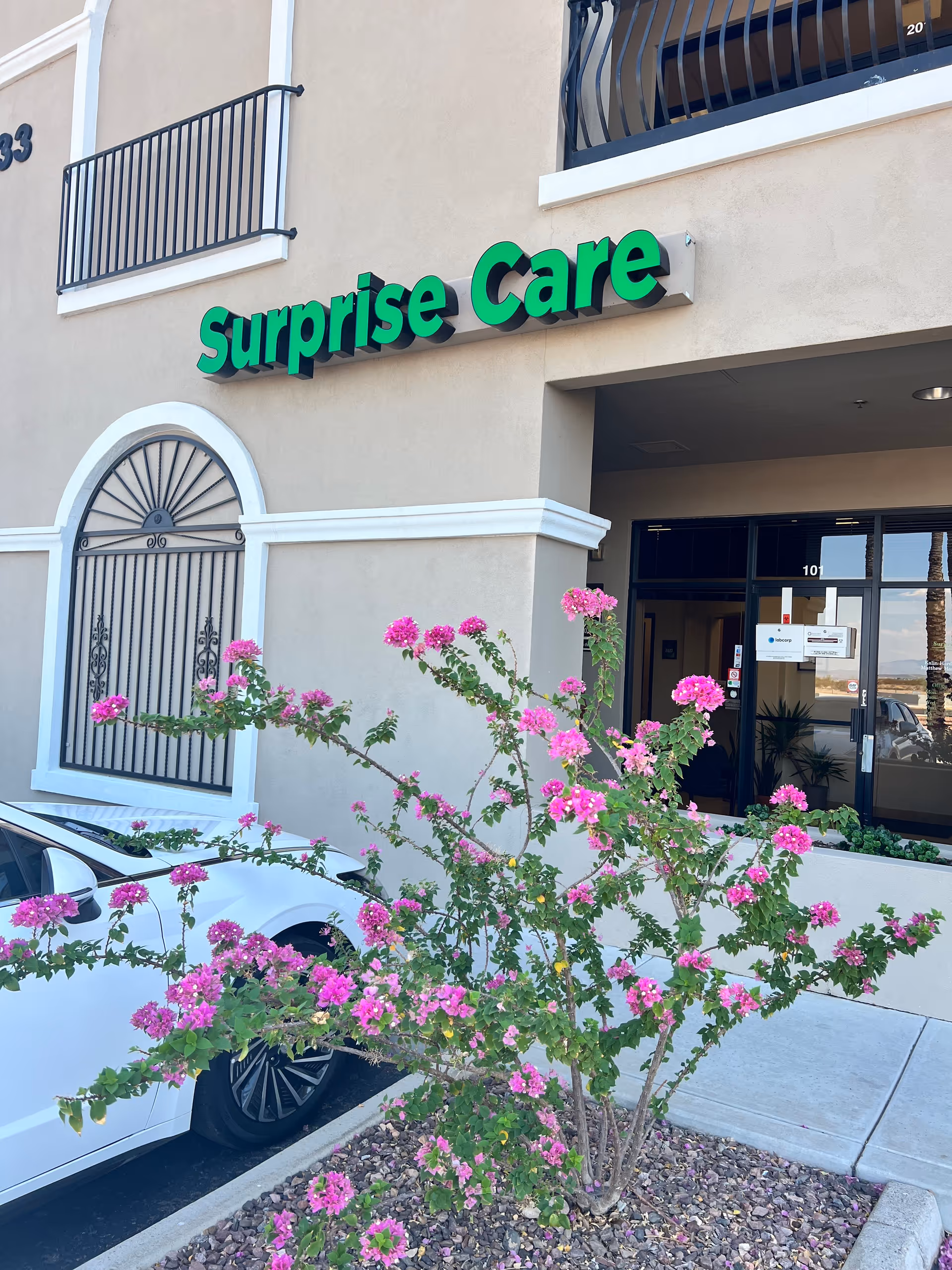 Exterior view of Surprise Care facility with green sign and flowering shrubs.
