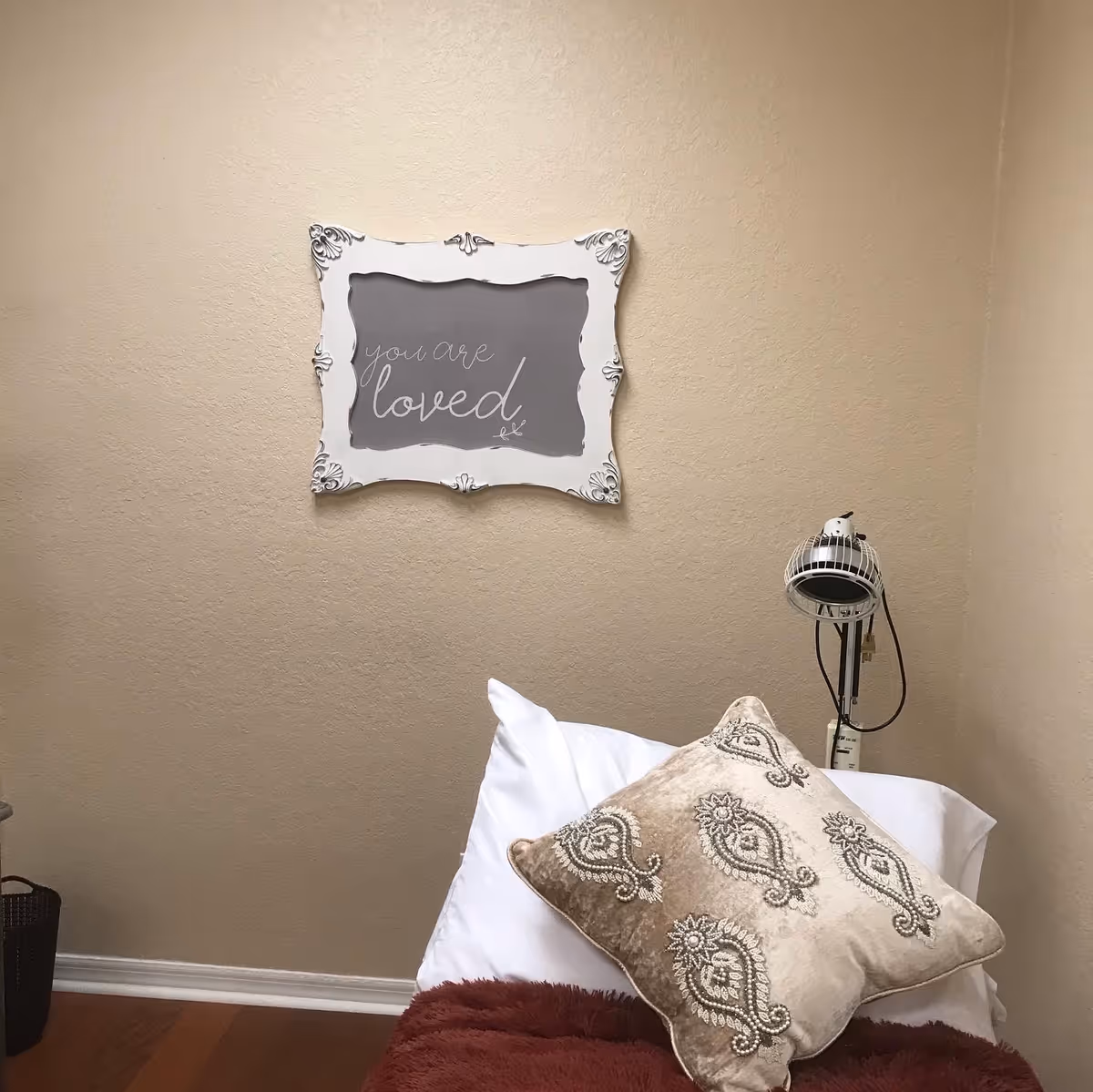 Cozy bedroom corner with a framed 'you are loved' sign, pillows, and a lamp.