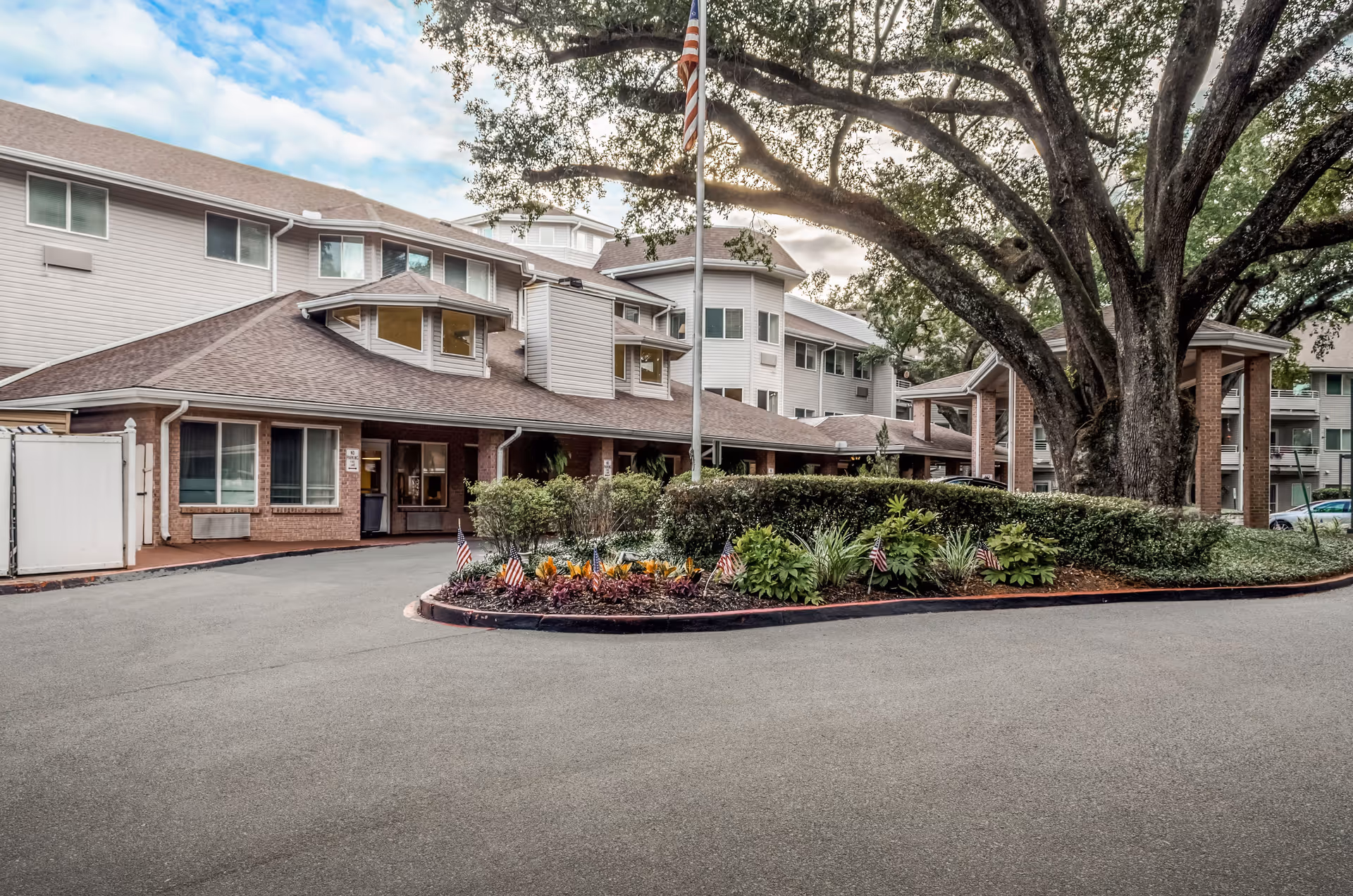 Exterior view of Asher Point Independent Living of Mobile, showing a multi-story building with beige siding and brick accents. There is a circular driveway with a landscaped island featuring small plants and American flags. A large tree stands prominently near the entrance, and an American flag flies on a flagpole in the center of the landscaped area.