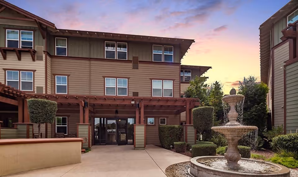 Courtyard entrance of a multi-story senior living building with a stone fountain and landscaped shrubs at sunset.