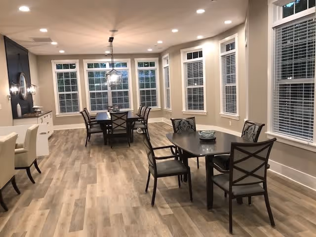 A spacious dining room with multiple dark wood tables and chairs arranged neatly on a light wood floor. The room features large windows with white blinds, beige walls, and recessed ceiling lights. A modern pendant light hangs above one of the tables, and there is a sideboard with decorative items along one wall.
