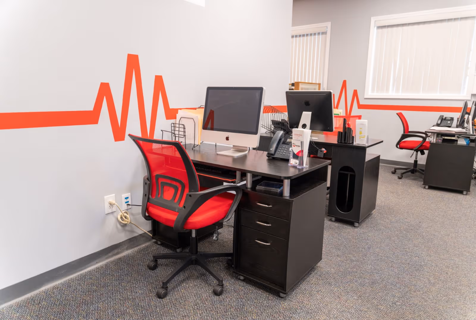 Office workspace with two desks, computers, and red chairs against a wall with a heartbeat mural.