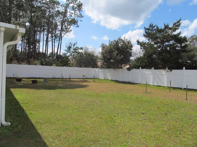 Green backyard with white fence, trees, and blue sky behind a house.