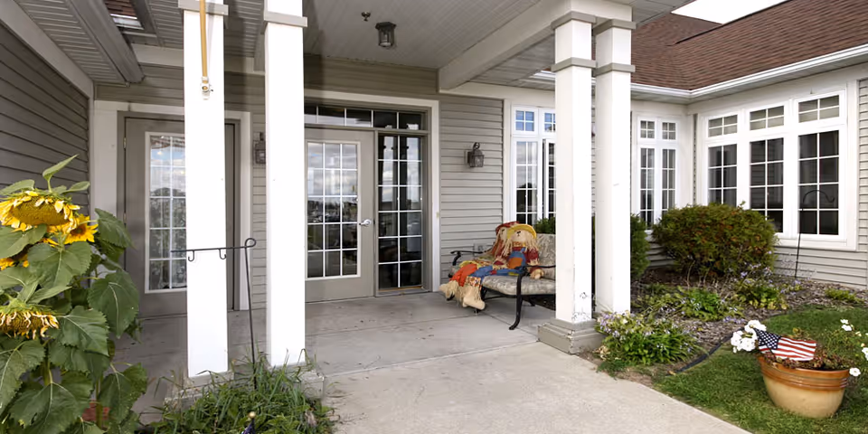 Covered front entrance with columns, glass doors, a bench holding a decorative scarecrow, and nearby plants and potted flowers.