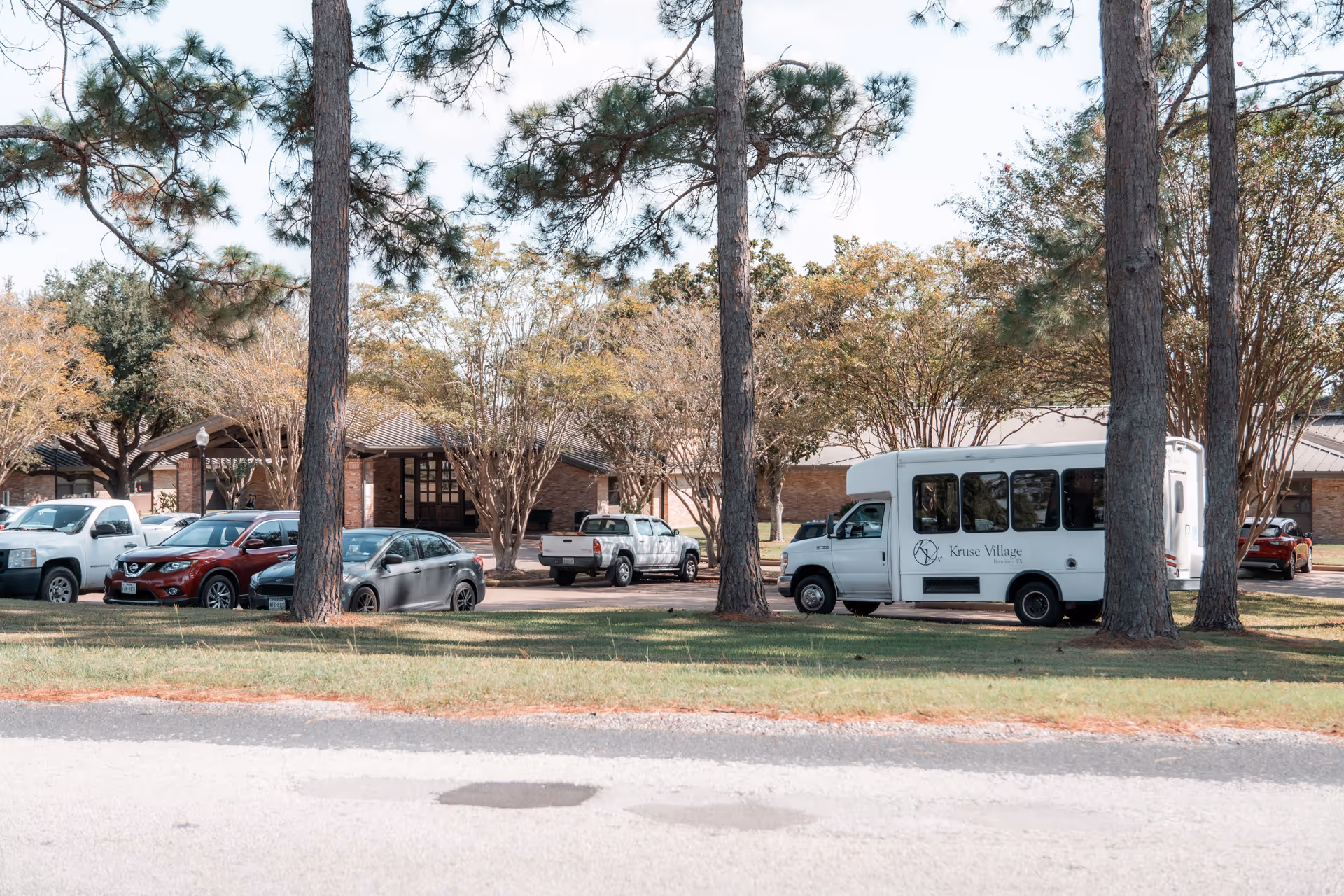 Front lawn and parking area of Kruse Village showing trees, several parked cars and a white Kruse Village shuttle bus in front of a low brick building.